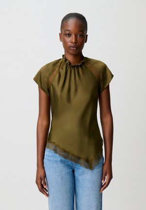 Donna che indossa una camicetta verde oliva a maniche corte con rifiniture in pizzo e jeans azzurri chiari, in piedi contro uno sfondo chiaro e semplice.