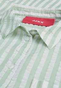 Camisa de rayas verde claro hecha de tela suave, con cuello, botones frontales y una etiqueta roja prominente con texto blanco.