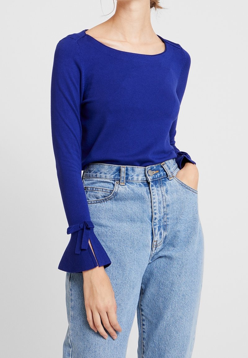 Top azul de manga larga con escote cuadrado; presenta mangas en forma de campana con volantes y detalles de lazo. Combinado con jeans de tiro alto y desgastados.