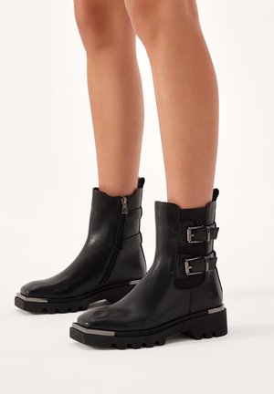 Black leather ankle boots with a slightly scalloped top, side zipper, and two buckle accents. Chunky rubber sole with metallic detailing.