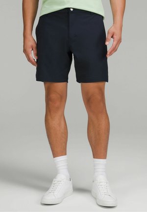 Homme portant un short noir, des chaussettes blanches montantes et des baskets blanches, debout devant un fond gris uni.
