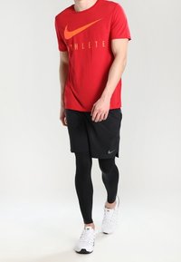 Rotes Nike-T-Shirt mit orangefarbenem Logo und dem Text "ATHLETE", kombiniert mit schwarzen Shorts und schwarzen Kompressionsleggings, abgerundet mit weißen Sneakers.