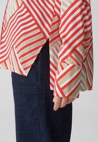 Camicia a righe caratterizzata da motivi alternati di rosso, beige e bianco, con una vestibilità ampia, maniche lunghe e dettagli laterali a spacco. Jeans in denim visibili.