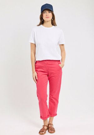 Femme portant un t-shirt blanc, un pantalon corail roulé aux chevilles, des sandales marron et une casquette de baseball bleue, debout devant un fond uni.