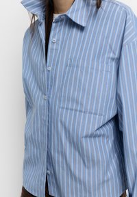 Camicia a righe blu chiaro con strisce verticali in blu scuro e arancione, chiusura con bottoni e una tasca sul petto.