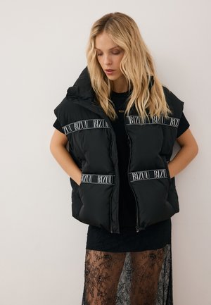 Blonde vrouw die een zwarte donzen vest draagt met de tekst "BIZUU", een zwart topje en een zwarte kanten rok, staand tegen een effen lichte achtergrond.