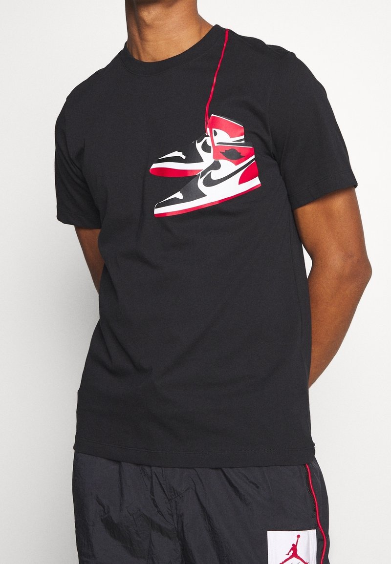 Svart t-shirt med kort ärm, prydd med en djärv grafisk bild av röda och svarta sneakers på framsidan. Den har rund halsringning och en mjuk tygtextur.