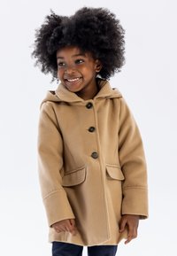 Manteau en polaire beige avec capuche, doté d'une fermeture à boutons, de deux poches avant et d'une coupe décontractée. Le manteau a une texture douce.