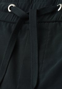 Gros plan d’un pantalon en tissu noir avec un cordon de taille passé à travers des œillets métalliques argentés et noué avec un nœud simple.