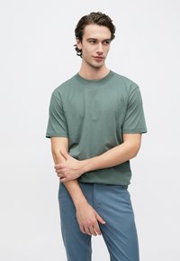 Πράσινο βαμβακερό t-shirt με κοντά μανίκια, στρογγυλή λαιμόκοψη και άνετη εφαρμογή, συνδυασμένο με ανοιχτό μπλε παντελόνι. Ομαλή υφή, μινιμαλιστικός σχεδιασμός.