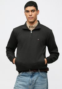 Pullover zip-up nero realizzato in tessuto morbido, con maniche lunghe e vestibilità rilassata. Piccolo logo sul petto, abbinato a jeans azzurri chiari.