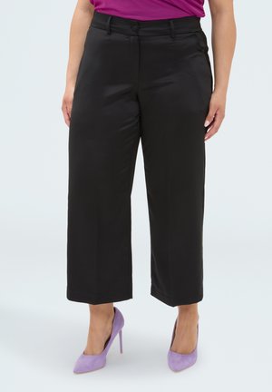 Donna con pantaloni neri a gamba larga cropped e scarpe con tacco alto color lavanda, in piedi su uno sfondo chiaro e uniforme.
