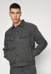 Σκοτεινό γκρι τζάκετ από τζιν με δύο τσέπες στο στήθος, κουμπιά μπροστά και δομημένο γιακά. Το ύφασμα έχει ελαφρώς υφασμένη επιφάνεια.