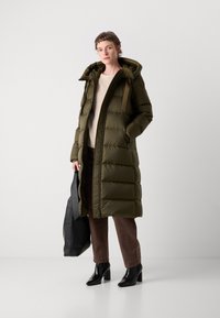 Abrigo acolchado verde oliva con capucha, diseño acolchado y cierre frontal. Combinado con un suéter crema, pantalones marrones y botas negras de tacón.