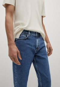 Persona vestida con jeans de mezclilla azul y una camisa de manga corta color crema, de pie con las manos relajadas a los lados.