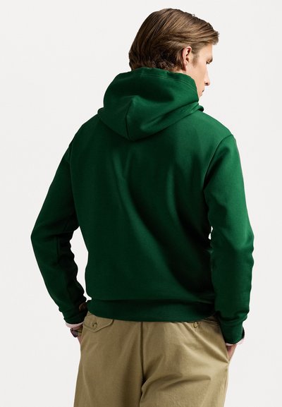 Green hoodie with a drawstring hood, soft texture, ribbed cuffs, and hem. Worn with khaki trousers, showing an overall casual design.