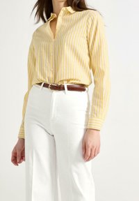 Gul- og hvidstribet skjorte med knapper og krave, parret med højtaljede hvide bukser og et brunt læderbælte.