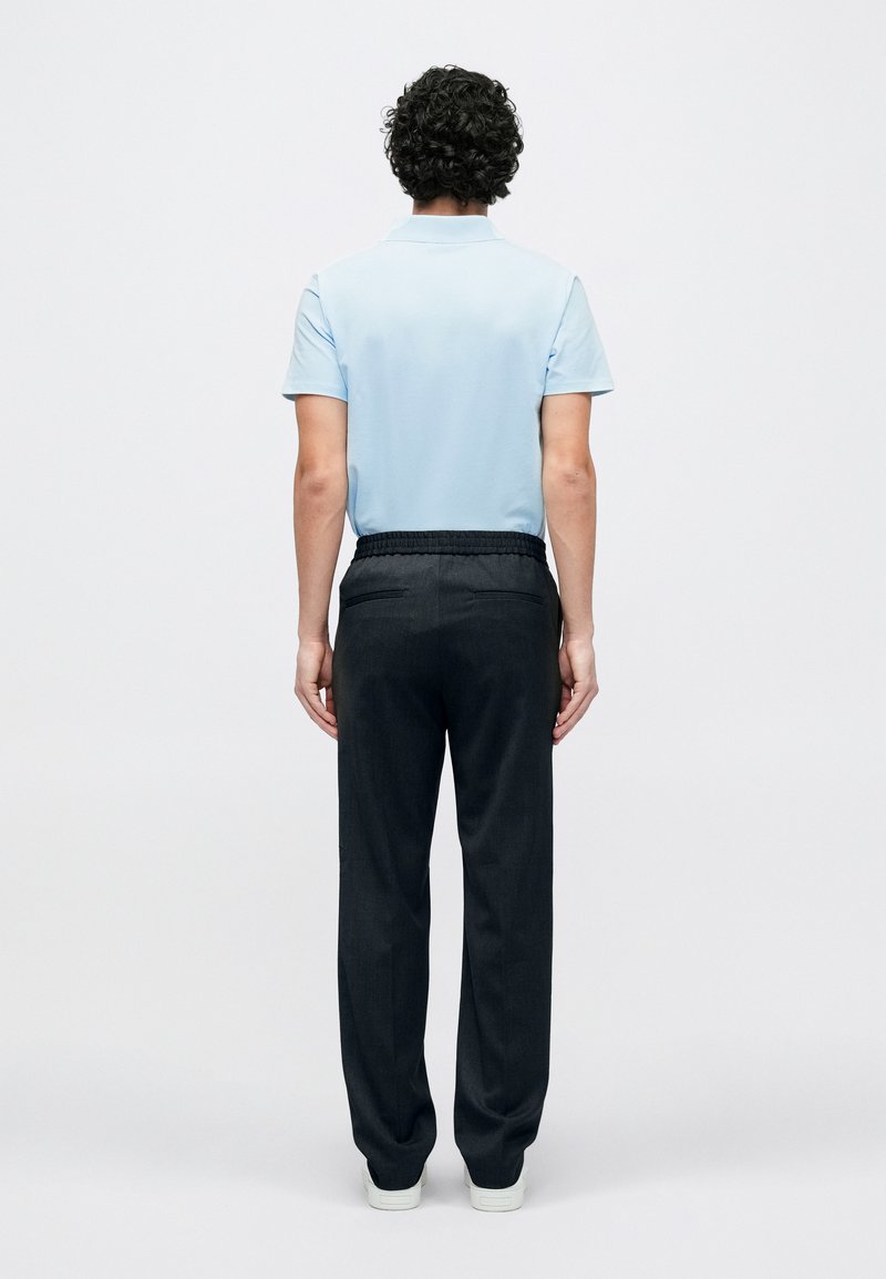 Hellblaues Polohemd mit kurzen Ärmeln, kombiniert mit dunkelgrauen Hosen mit elastischem Bund. Modell blickt nach hinten, minimalistisch gestalteter Hintergrund.