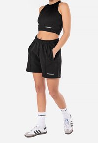 Osoba v černém bezrukávovém crop topu a černých šortkách s kapsami, bílých ponožkách a bílých teniskách s černými pruhy.