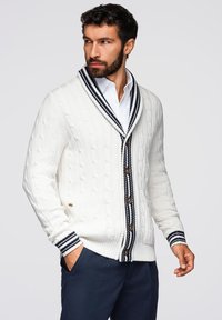 Cardigan bianco a maglia spessa con colletto a scialle, dettagli a righe blu navy e bianche, bottoni in legno e tasche frontali. Motivo di maglia testurizzato.
