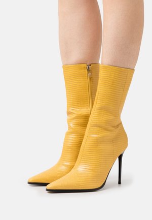 Bottes à talons hauts texturées jaune montant jusqu'au milieu du mollet, avec des bouts pointus et des fermetures éclair latérales, portées sur des jambes nues sur un fond clair uni.