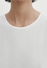 Haut blanc à manches courtes en tissu texturé et respirant, avec un col rond et un détail de bordure contrastante.