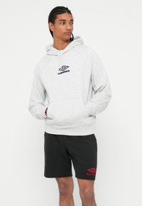 Giovane con capelli intrecciati, indossa una felpa leggera grigia e pantaloni corti neri, entrambi con il logo Umbro, in posa con le mani nella tasca della felpa.