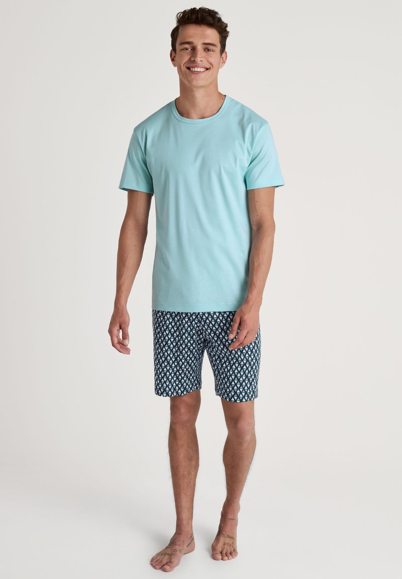 Lichtblauw katoenen T-shirt met ronde hals, gecombineerd met marineblauwe zwembroek met een patroon van witte geometrische vormen. Naaktvoets model staat.