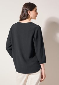 Svart topp med en texturerad, kramad design, 3/4 långa ärmar och rundad nederdel. Har en vid halsringning och en något avslappnad passform.