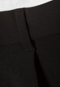 Gros plan sur un pantalon en tissu noir avec une boucle de ceinture et une partie d'une ceinture blanche visible en haut.