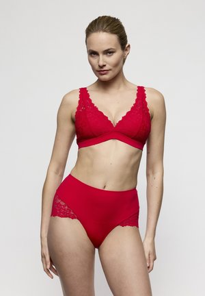 Vrouw die een rode kanten beha en een hoge taille slip draagt, staand tegen een effen witte achtergrond met haar handen ontspannen langs haar lichaam.