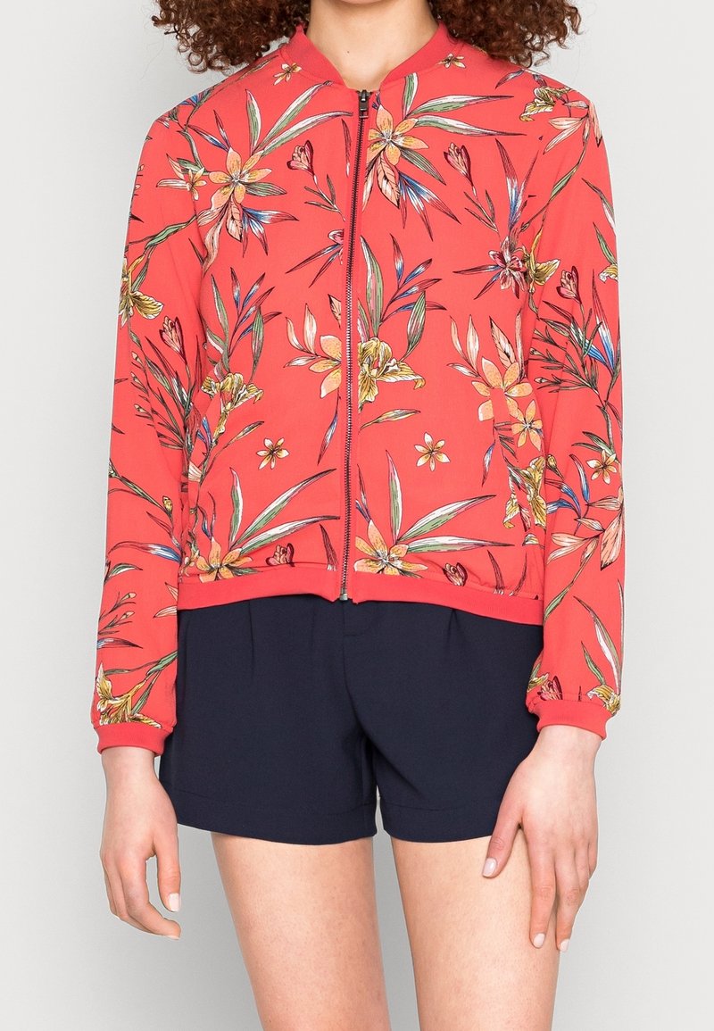 Veste bomber rouge à motif floral, fermeture zippée et poignets côtelés. Présente des fleurs et des feuilles colorées, portée avec un short bleu marine.
