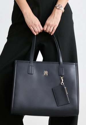 Personne tenant un sac cabas en cuir noir avec deux poignées et une petite pochette attachée, portant une tenue noire et une montre-bracelet.