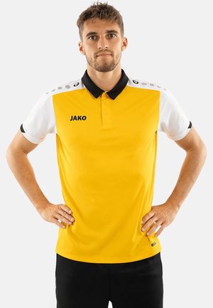 Junger Mann steht mit den Händen in den Hüften, trägt ein gelb-weißes Polohemd mit schwarzem Kragen und schwarze Hose vor einem einfarbigen Hintergrund.
