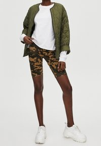 Gequilte bomberjack in olijfgroen gedragen over een wit shirt met lange mouwen, gecombineerd met bruine camouflage biker shorts en witte sneakers.