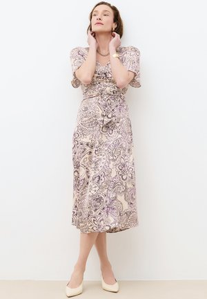 Frau trägt ein kurzärmeliges Kleid mit kurzem gesäumtem Midirock in beige und lila Paisley-Muster, eine goldene Kettenkette, ein Armband und cremefarbene Absätze.