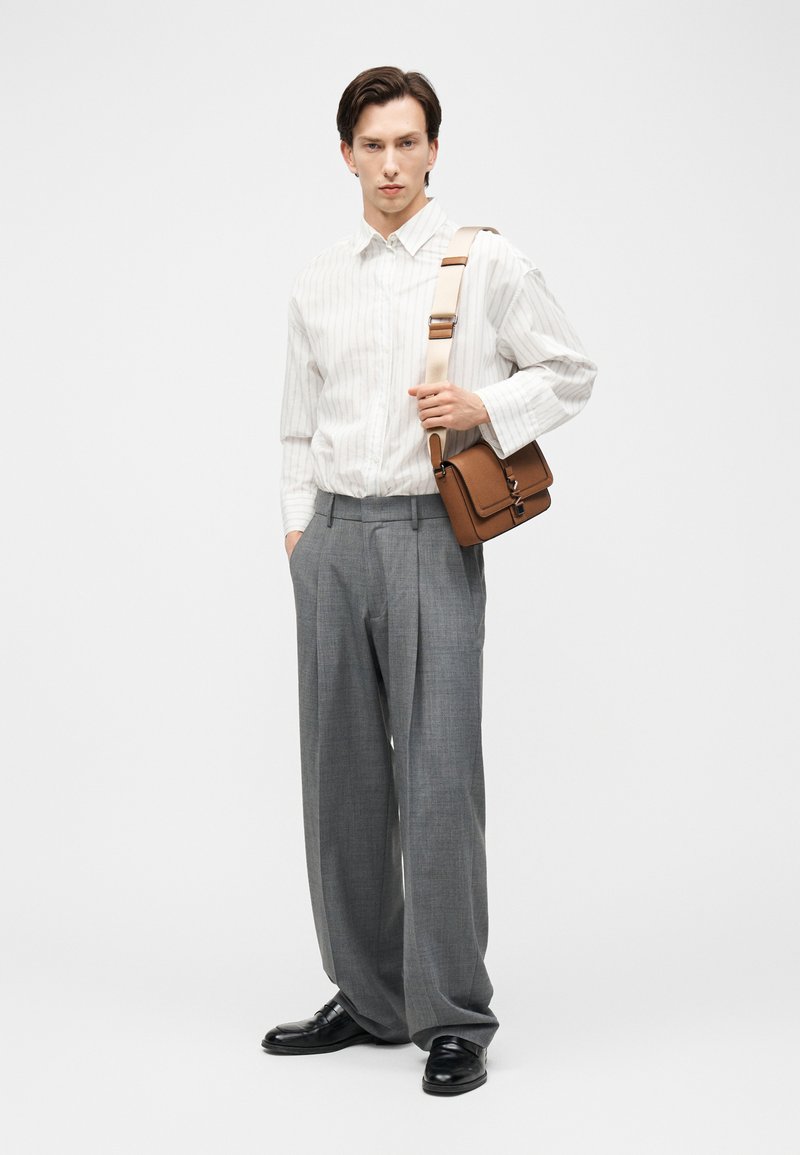 Joven con camisa blanca de rayas, pantalones grises de pierna ancha, zapatos negros, sosteniendo un bolso marrón con correa beige sobre fondo blanco.