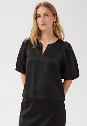 Femme blonde portant un chemisier en satin noir avec des manches courtes bouffantes et une encolure en V, posant devant un fond clair uni.