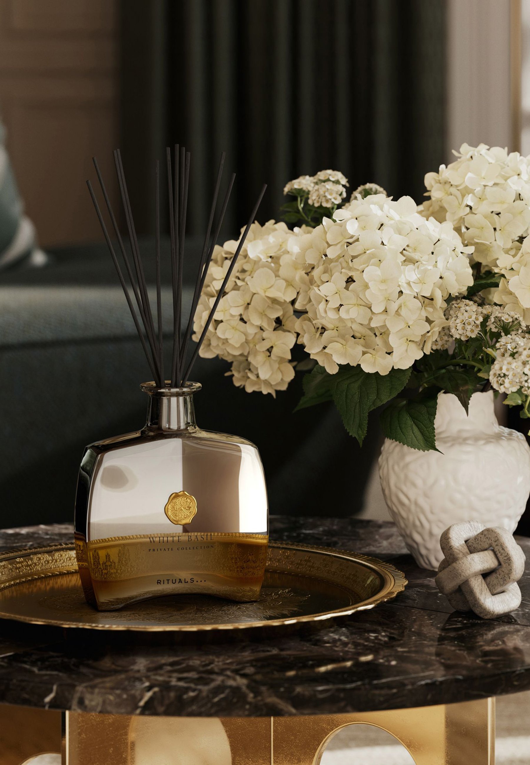 Reed diffuser bottle in transparent glass with metallic accents, featuring black reeds and a gold label. Includes a white floral arrangement.
