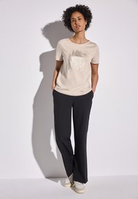 T-shirt beige avec un design graphique au centre, associé à un pantalon noir ample et des baskets blanches. Tissu lisse avec une texture douce.
