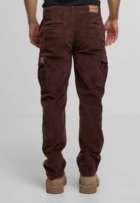 Mies, joka on pukeutunut tummanruskeisiin korvakuulokkeisiin ja beigeihin saappaisiin, seisoo selkä kameraan päin vaaleaa taustaa vasten.