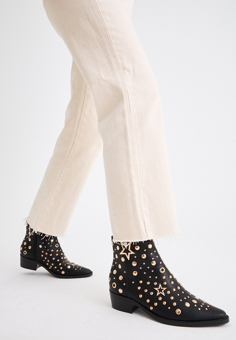 Botines negros a los tobillos con tachuelas doradas y acentos de estrellas, punta afilada, tacón bajo en bloque y superficie de cuero texturizado, combinados con jeans de color claro.