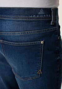 Vaqueros de mezclilla azul oscura con un bolsillo trasero, costuras doradas y un sutil logo en forma de triángulo cerca de la cinturilla. Textura suave, diseño clásico.
