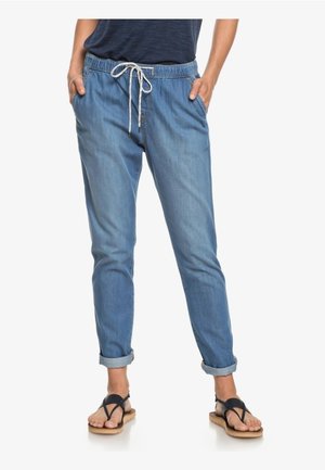 Vrouw draagt lichtblauwe denim joggerbroek met omgeslagen pijpen en wit trekkoord, een donkerblauw topje en zwarte sandalen, staand met handen in de zakken.