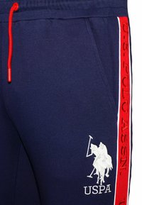 Navy sweatpants with red drawstring and side stripes. Features white embroidered logo of a polo player. Soft fabric with a smooth texture.