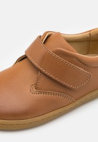 Zapato de cuero color marrón claro con una amplia tira de velcro, forma redondeada, textura suave y costuras en contraste. Tiene una suela flexible para mayor comodidad.