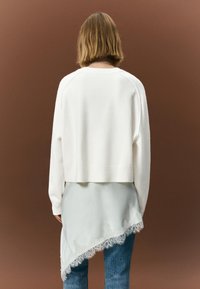 Biely skrátený sveter s dlhými rukávmi vrstvený cez svetlomodrý asymetrický top s čipkovanou ozdobou, doplnený modrými džínsami.