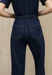 Mono de mezclilla índigo con mangas cortas, cintura ajustada y cuatro bolsillos. Presenta costuras en contraste y una textura suave y ligera.