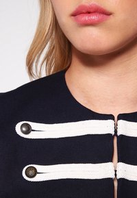 Granatowy top z tkaniny z warkoczowymi białymi akcentami i srebrnymi guzikami, z wysokim dekoltem i detalem w formie frontowego zapięcia.