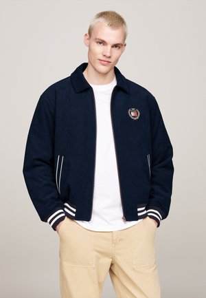 Giacca navy con zip, polsini a costine e strisce bianche. Presenta un logo sul lato sinistro del petto. Abbinata a una t-shirt bianca e pantaloni beige.
